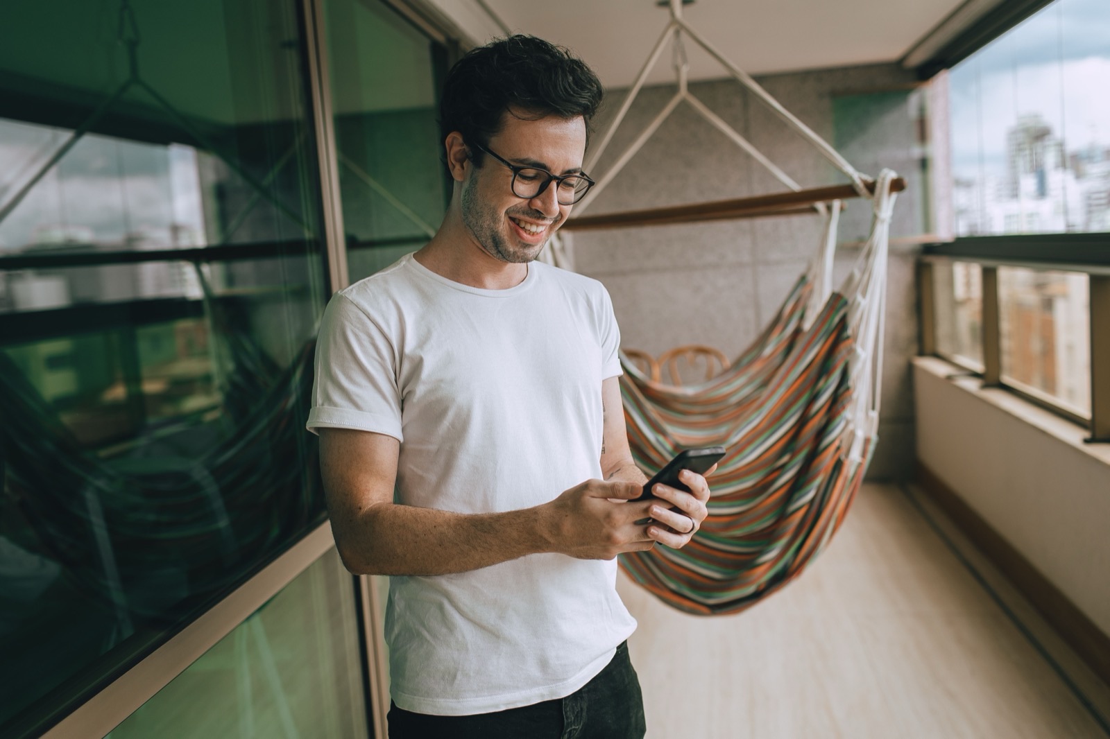 A traveller stands on a city balcony, smiling at their phone, with a hammock chair and rooftops behind them.